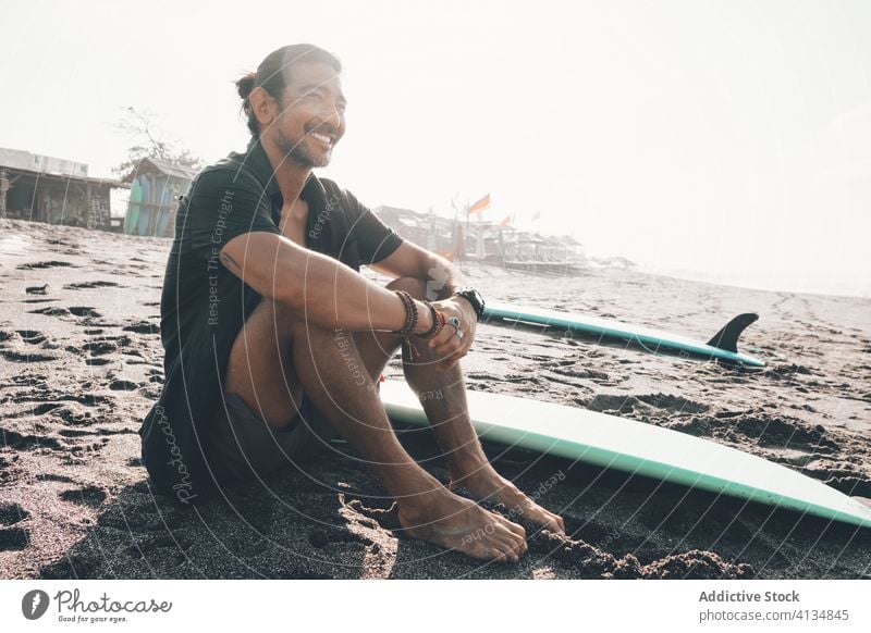 Glücklicher ethnischer Mann mit Surfbrett am Strand sitzend positiv ruhen MEER genießen Sand Sommer jung hispanisch männlich Lifestyle Urlaub Küste Meer Ufer