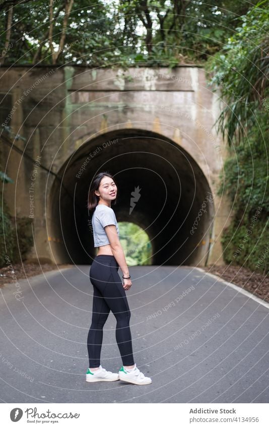 Schlanke asiatische Frau steht auf einer asphaltierten Straße in der Nähe eines Tunnels Stollen Asphalt Baum Pflanze Sportbekleidung Wochenende Erholung
