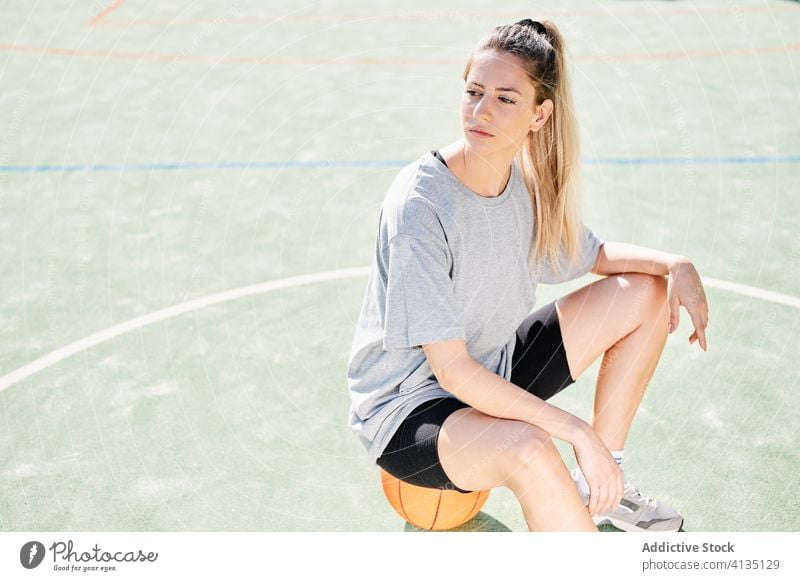 Sportliche Frau sitzt auf einem Basketball auf dem Platz selbstbewusst Gericht sitzen ruhen Ball ernst jung Sportlerin Athlet Sportbekleidung Training Aktivität