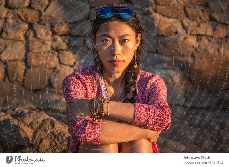 Junge ethnische Frau sitzt an einer verwitterten Steinmauer jung Windstille Porträt Steinwand antik sitzen sich[Akk] entspannen asiatisch Stil lässig trendy