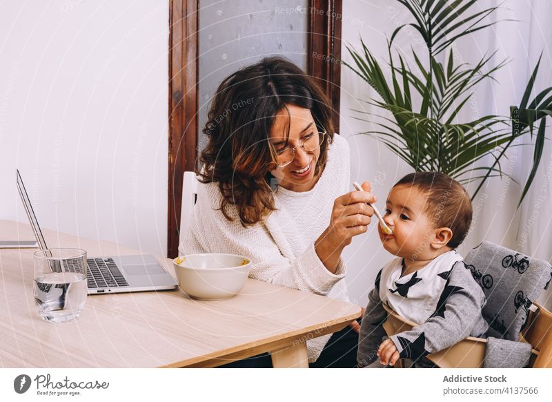Vielbeschäftigte Mutter, die ihr Kind füttert und von zu Hause aus arbeitet Frau Arbeit heimwärts Futter Laptop modern Anschluss interagieren jung Baby Gerät