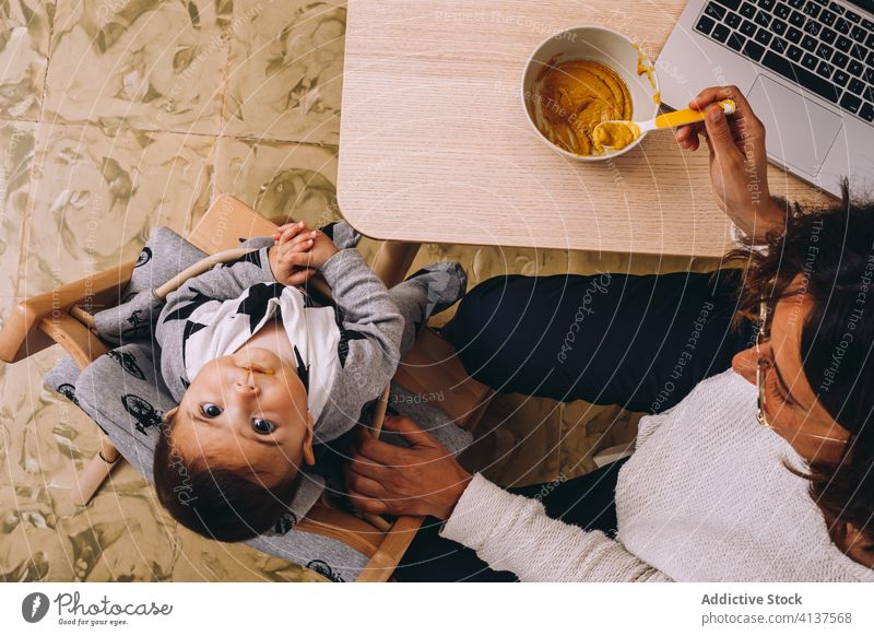 Vielbeschäftigte Mutter, die ihr Kind füttert und von zu Hause aus arbeitet Frau Arbeit heimwärts Futter Laptop modern Anschluss interagieren jung Baby Gerät