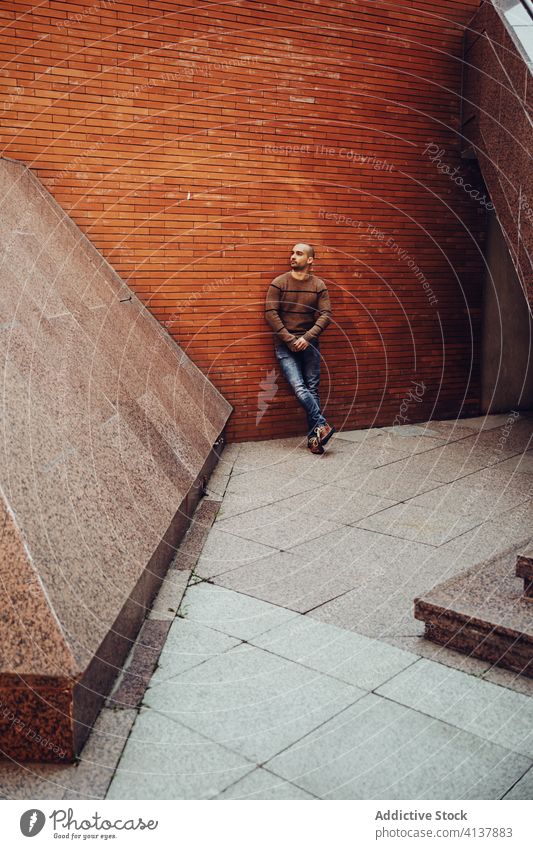 Stilvoller junger Mann steht neben Gebäude selbstbewusst lässig modern kahl Wand urban Straße männlich Trägerkleid Sweatshirt Jeanshose trendy Typ Zeitgenosse