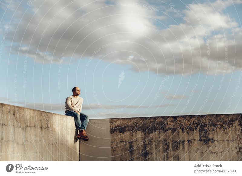 Junger Mann sitzt auf einer Betonkonstruktion auf der Straße Zaun Stein schäbig Konstruktion urban allein Hipster modern Himmel Wolken sonnig Typ sitzen lässig
