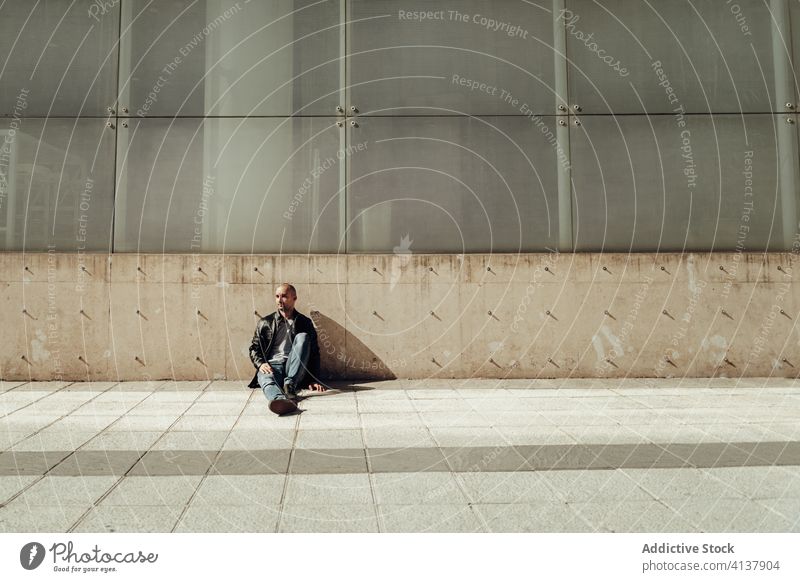 Ethnischer Mann in trendigem Outfit sitzt auf der Straße Hipster jung Stil trendy Gebäude Zeitgenosse Porträt modern Typ lässig zerrissen Jeanshose Vollbart