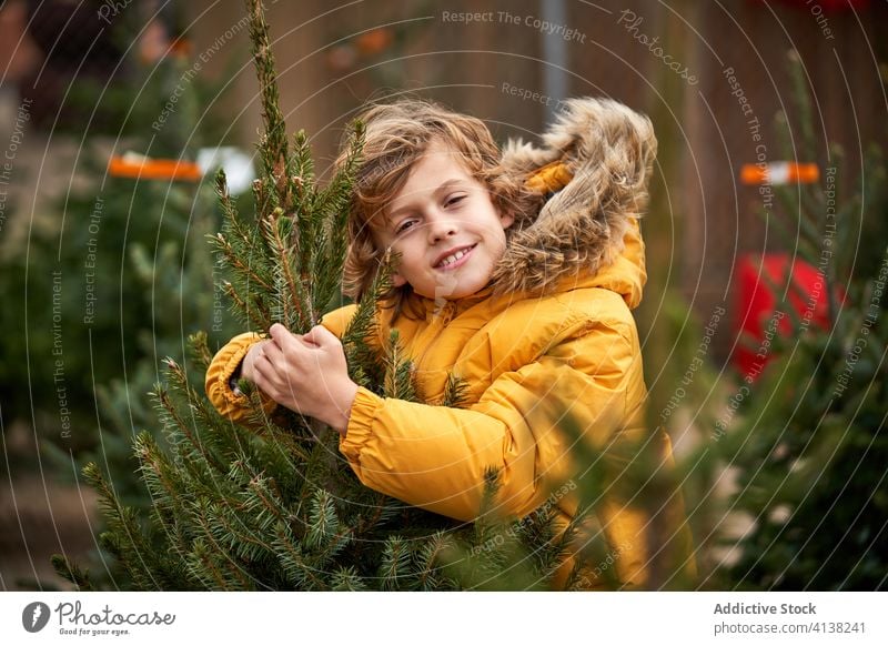 Schöner blonder Junge mit gelber Jacke kauft seinen Weihnachtsbaum Lächeln finden außerhalb Kiefer verschneite Jungen Kindheit laufen saisonbedingt jung Jugend