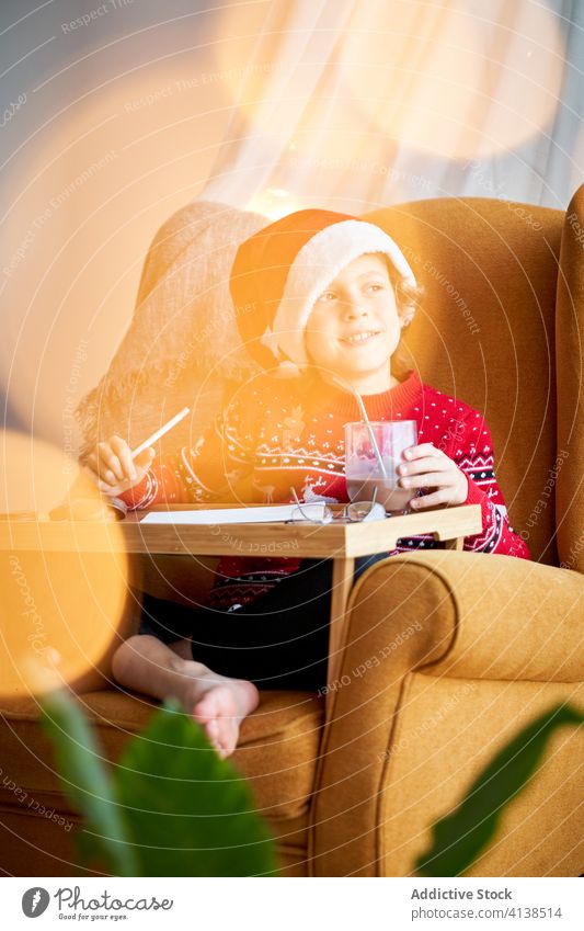 Fröhlicher Junge in Weihnachtskleidung im gemütlichen Sessel sitzend Weihnachtsmütze Weihnachten Feiertag Bleistift Armsessel Glück Komfort heimisch Pullover