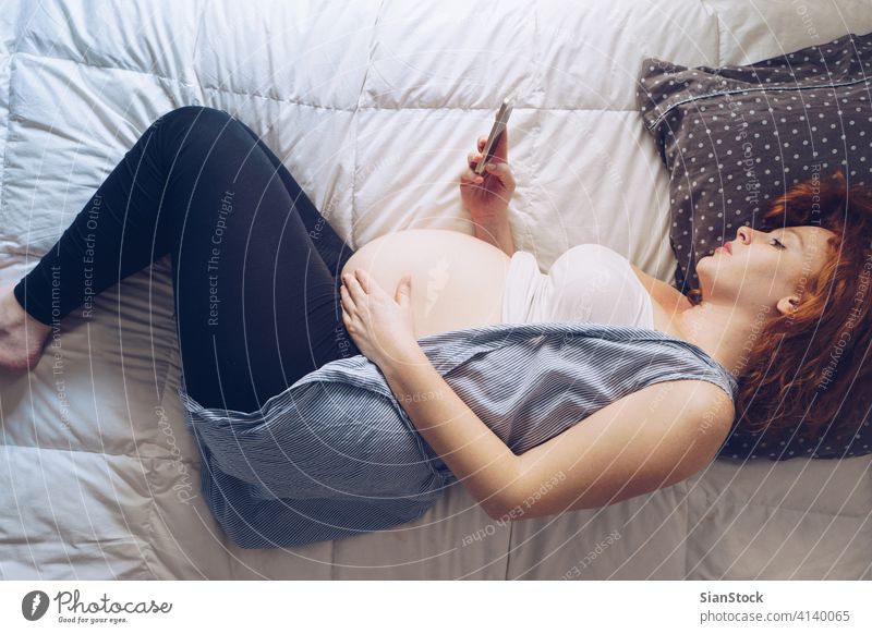 Schwangere Frau liegt mit Smartphone im Bett, Draufsicht schwanger Schwangerschaft Morgen jung Mädchen Sommersprossen Kopfkissen Sonntag entspannend