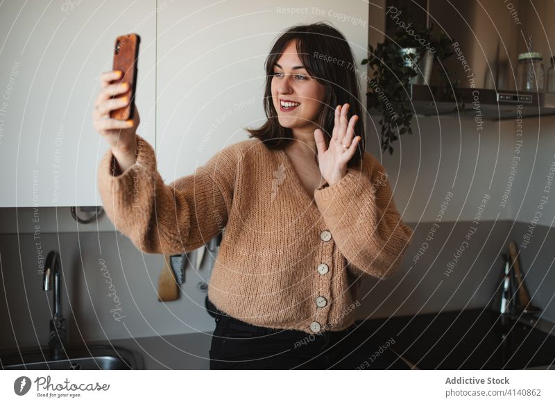 Fröhliche Frau, die sich zu Hause per Smartphone über einen Videochat unterhält Videoanruf Gespräch Video-Chat gestikulieren Gruß Wellenhand benutzend heimwärts