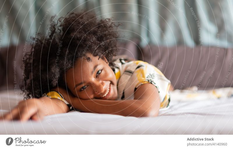 Fröhliche afroamerikanische Frau ruht sich auf dem Bett in einer modernen Wohnung aus Kälte heimwärts Lügen Lächeln heiter ruhen Appartement gemütlich ethnisch