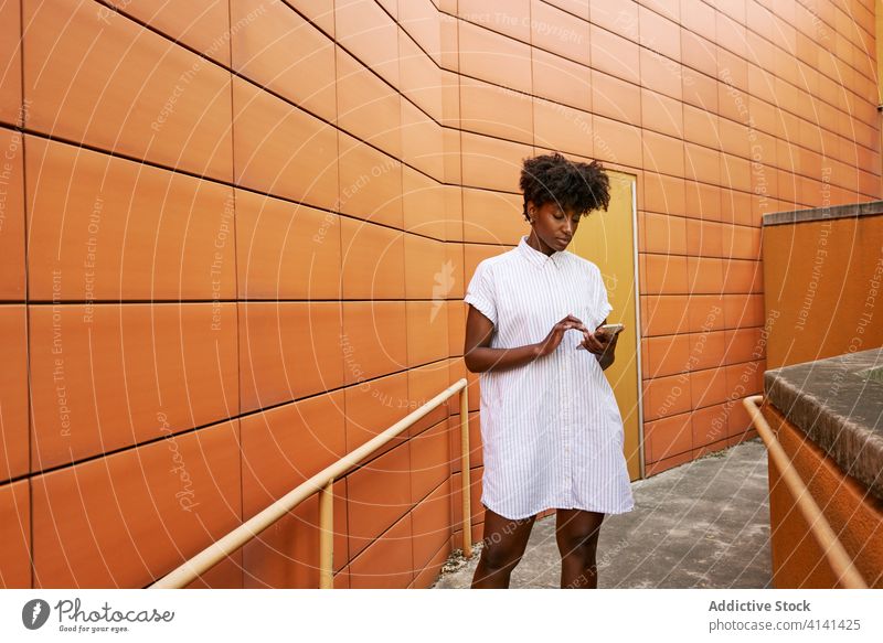 Fokussierte schwarze Frau mit Smartphone auf der Straße gegen lebhaftes Äußeres eines modernen Gebäudes Nachrichtenübermittlung Weg Kälte Browsen verwenden