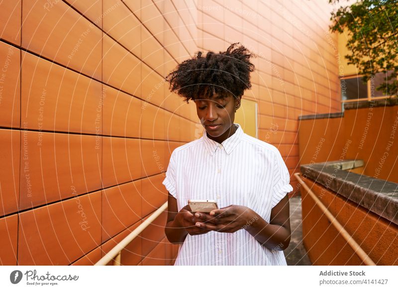 Fokussierte schwarze Frau mit Smartphone auf der Straße gegen lebhaftes Äußeres eines modernen Gebäudes Nachrichtenübermittlung Weg Kälte Browsen verwenden