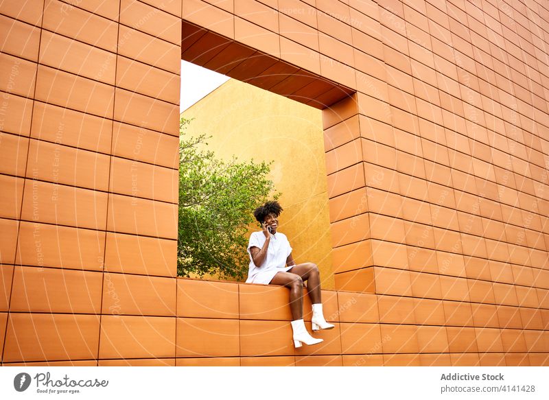 Stilvolle freudige schwarze Frau spricht am Telefon auf der Straße in der Nähe von Gebäude mit bunten außen Smartphone Anruf Architektur Außenseite urban Park