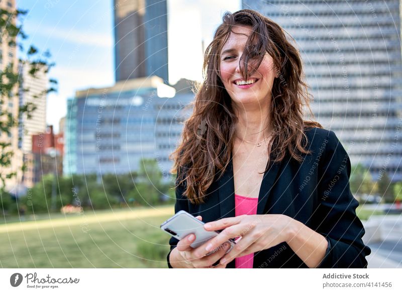 Fröhliche Geschäftsfrau in Jacke, die auf der Straße ihr Smartphone durchsucht Unternehmer benutzend Großstadt stilvoll Browsen Lächeln urban Frau Mobile