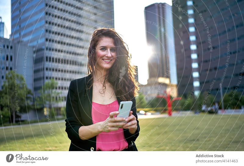 Fröhliche Geschäftsfrau in Jacke, die auf der Straße ihr Smartphone durchsucht Unternehmer benutzend Großstadt stilvoll Browsen Lächeln urban Frau Mobile