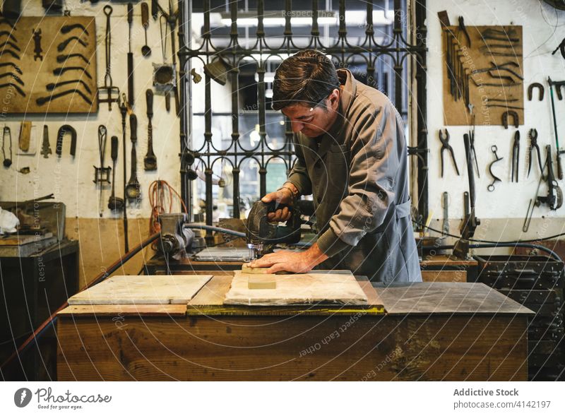 Ethnischer Juwelier mit Bohrmaschine bei der Arbeit mit Holz in der Werkstatt bohren Klotz hölzern Basteln Uniform Konzentration Kunsthandwerker