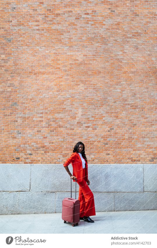 Schwarze Frau geht mit Koffer auf dem Bürgersteig Spaziergang Mode Glamour Stil Straßenbelag rot Backsteinwand Gebäude gestikulieren elegant farbenfroh