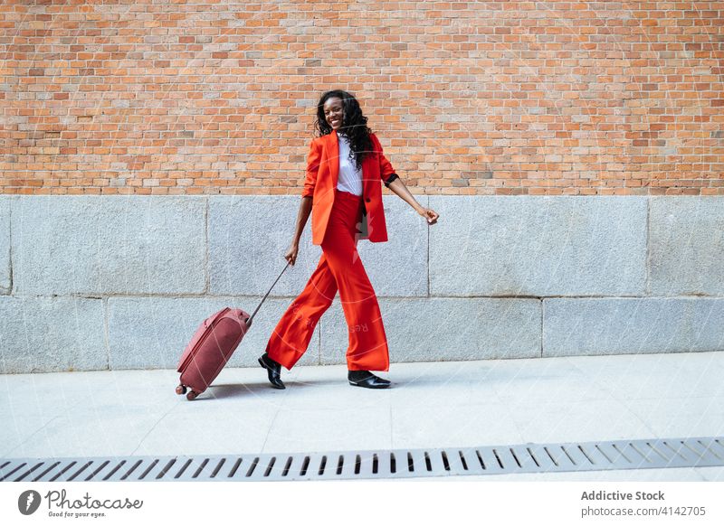 Schwarze Frau geht mit Koffer auf dem Bürgersteig Spaziergang Mode Glamour Stil Straßenbelag rot Backsteinwand Gebäude gestikulieren elegant farbenfroh