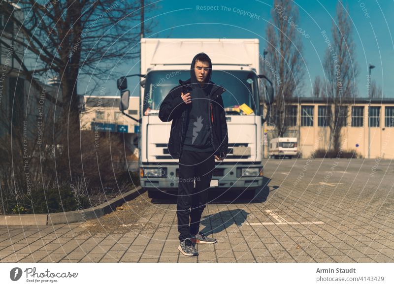 junger Mann mit Rucksack steht vor einem Lastwagen aeria Architektur schön schwarz Junge Gebäude lässig Kaukasier selbstbewusst Kapuzenpulli Lifestyle Blick