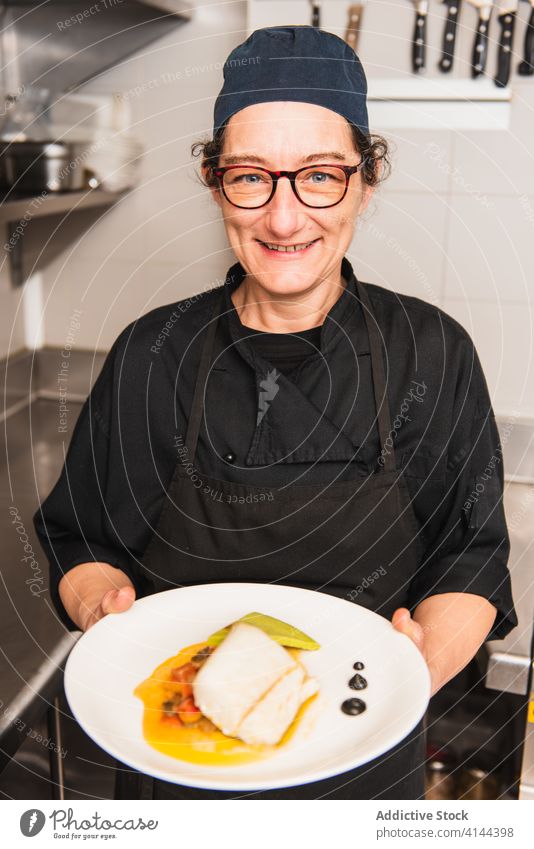 Porträt einer Köchin, die ein Kabeljau-Gericht hält. Speise Fisch Lebensmittel Küchenchef Restaurant Dorsch Feinschmecker Teller MEER Hut Catering Charakter