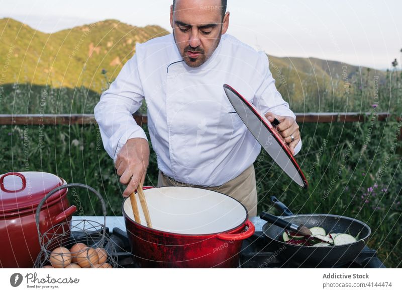 Professioneller Koch bei der Zubereitung von Speisen am Herd in der Natur Mann Kochtopf Berge u. Gebirge malerisch Küchenchef vorbereiten Lebensmittel lecker
