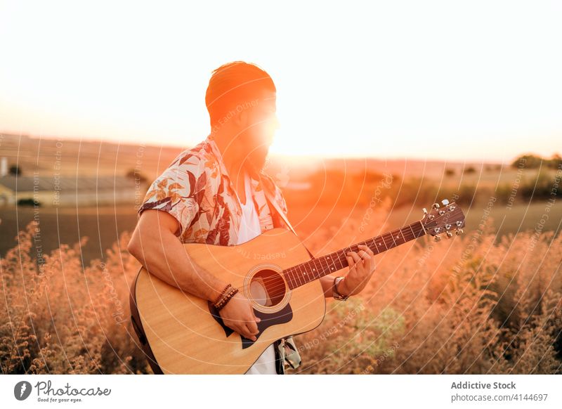 Männlicher Musiker spielt Gitarre auf dem Land bei Sonnenuntergang spielen Hipster Gras Himmel Feld Harmonie Mann Musical Instrument Gitarrenspieler künstlich