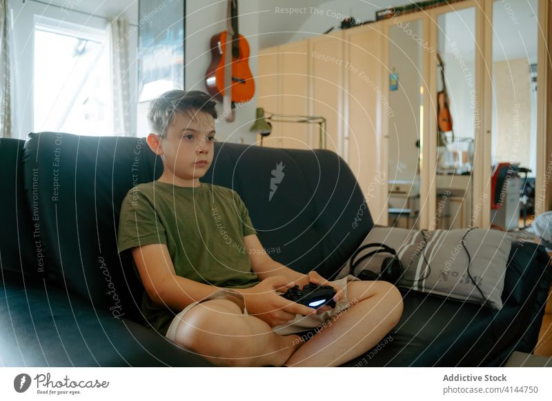Konzentriertes Kind beim Videospiel auf der Couch Junge spielen Joystick Sofa Beine gekreuzt heimwärts unterhalten Gerät Apparatur Freizeit Konzentration wenig