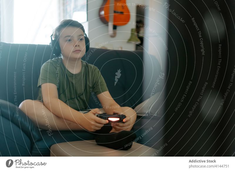 Konzentriertes Kind beim Videospiel auf der Couch Junge spielen Joystick Sofa Beine gekreuzt Kopfhörer heimwärts unterhalten Gerät Apparatur Freizeit