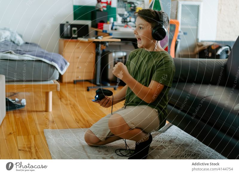 Fröhliches Kind hat Spaß beim Videospiel zu Hause Junge feiern Kopfhörer Joystick Sieg aufgeregt heiter spielen heimwärts unterhalten Gerät Glück wenig lässig