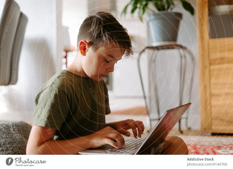 Fokussiertes Kind benutzt Laptop zu Hause Junge Tippen Stock heimwärts benutzend Internet ernst Beine gekreuzt Gerät Apparatur Browsen online Anschluss wenig