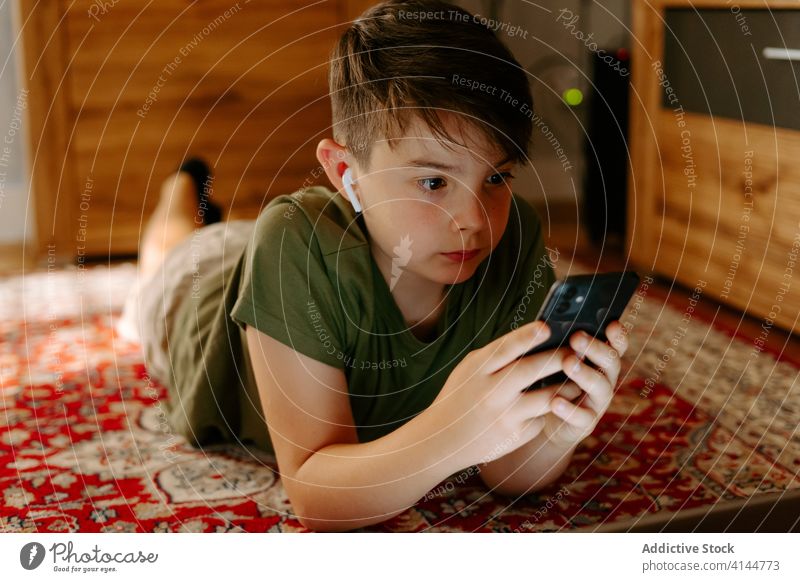 Fokussiertes Kind mit drahtlosen Ohrhörern und Smartphone zu Hause Junge zuschauen Lügen Teppich heimwärts Drahtlos Kopfhörer Konzentration Gerät Apparatur