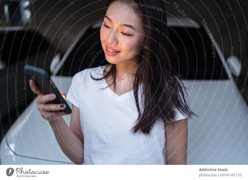 Glückliche asiatische Frau, die in der Nähe des Autos auf ihrem Smartphone surft benutzend Garage PKW Lächeln dunkel soziale Netzwerke Textnachricht Süchtige