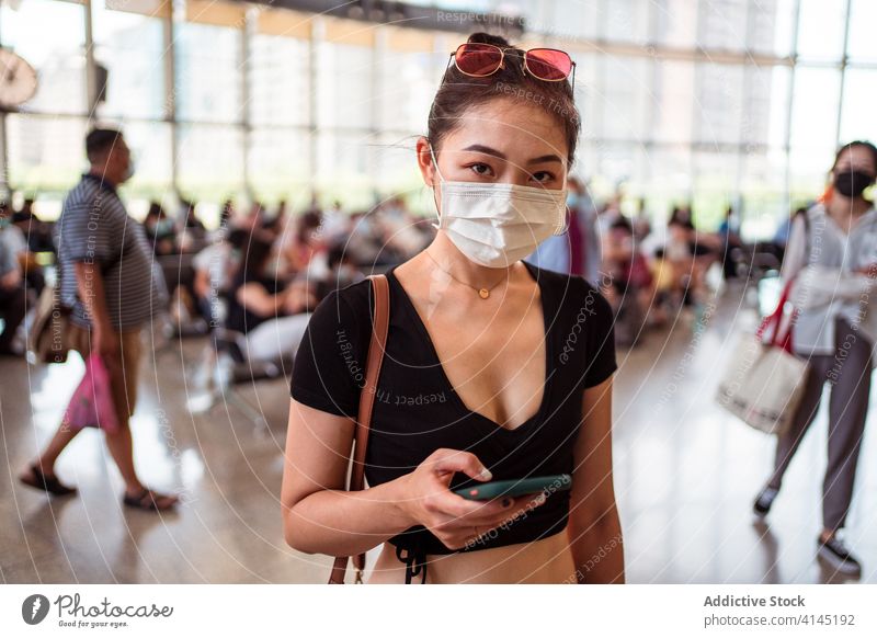 Asiatische Frau benutzt Smartphone in einem modernen Flughafen Abheben räkeln benutzend Mundschutz behüten Menge Coronavirus ethnisch asiatisch Zeitgenosse