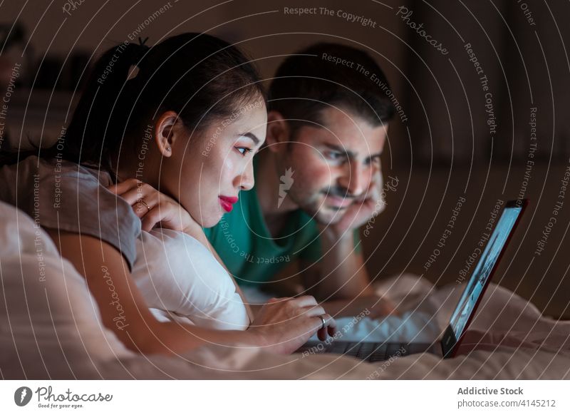 Glückliches multiethnisches Paar sieht sich im Bett einen Film auf dem Laptop an zuschauend teilen Komfort heimisch benutzend Apparatur Gerät Netbook Internet