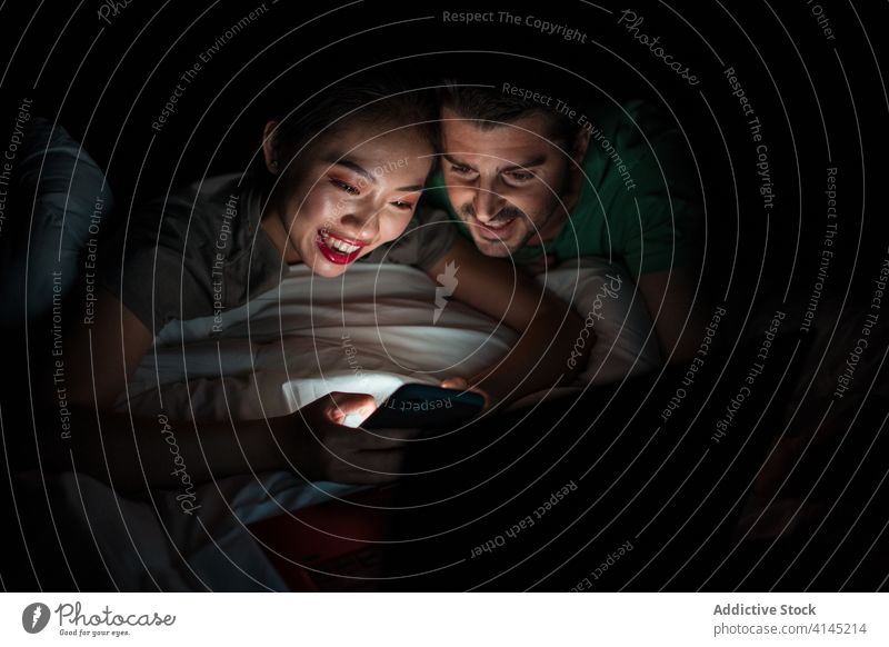 Ein gemischtrassiges Paar surft zu Hause im Bett mit seinem Smartphone im Internet soziale Netzwerke teilen Partnerschaft Surfen Nacht benutzend Apparatur Gerät