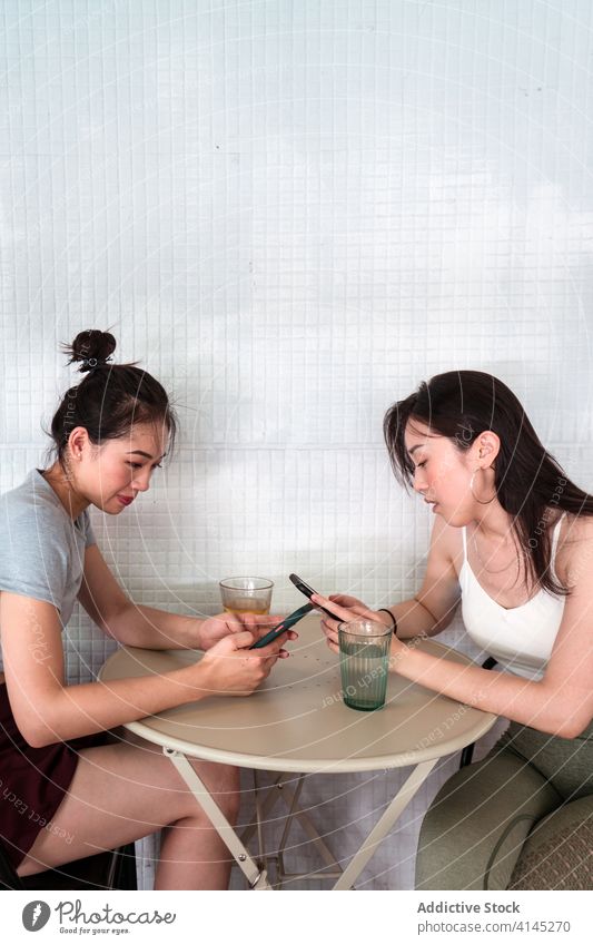 Asiatische Freundinnen, die in der Cafeteria mit ihren Smartphones im Internet surfen plaudernd soziale Netzwerke Surfen online Wasser Beine gekreuzt benutzend