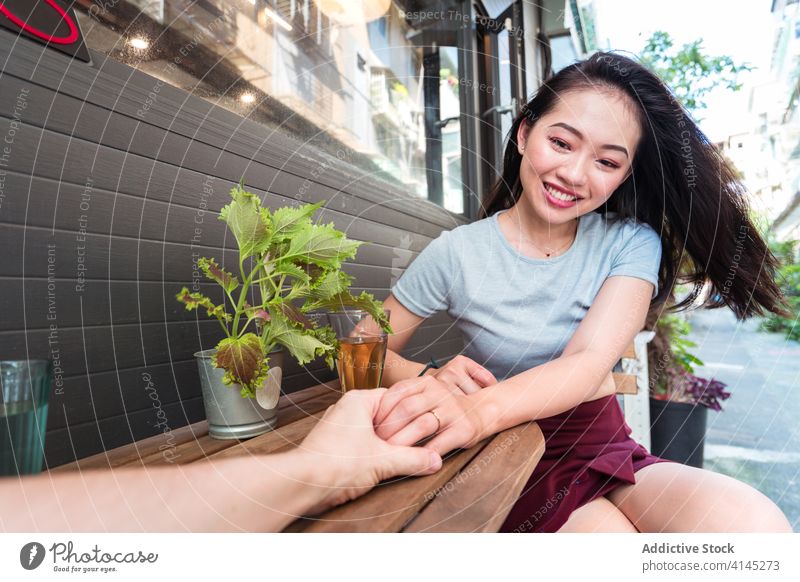 Fröhliche asiatische Frau im Gespräch mit einem nicht erkennbaren Freund in einem Cafe Kommunizieren Getränk Glas aufgeregt Lachen Zusammensein Freizeit