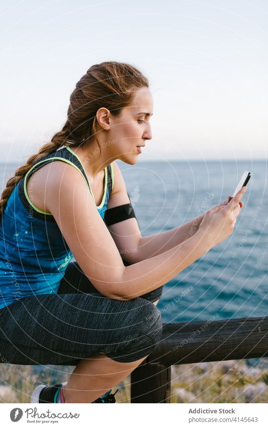 Lächelnde Frau in Sportkleidung mit Smartphone am Meer Sportlerin Stauanlage Browsen sich[Akk] entspannen Training MEER Sportbekleidung benutzend Fitness