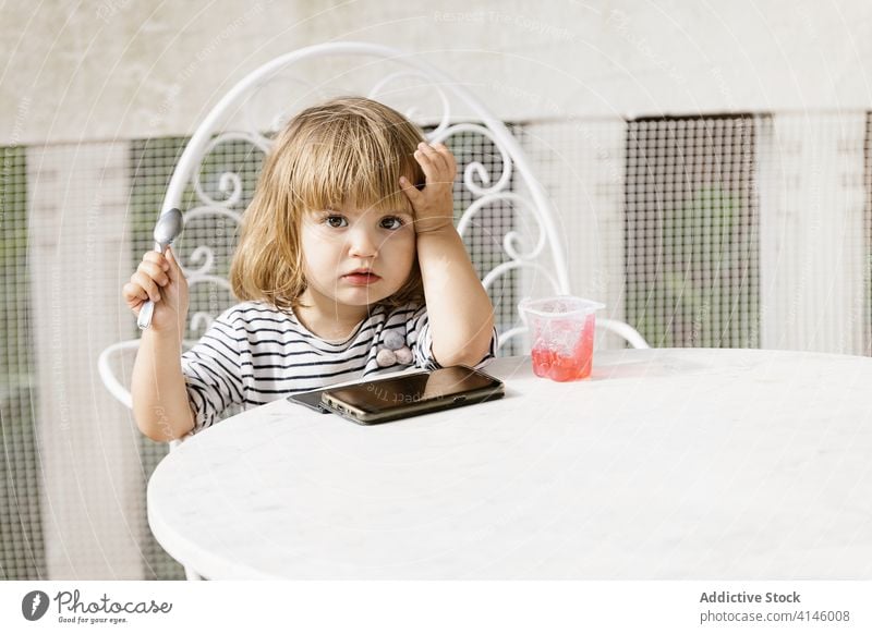 Kind isst Gelee und verwendet Smartphone auf Terrasse zuschauen Karikatur benutzend wenig Mädchen unterhalten Sommer Hinterhof Tisch niedlich Kindheit sitzen