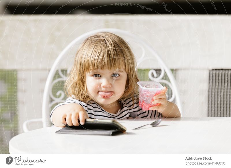 Kind isst Gelee und verwendet Smartphone auf Terrasse zuschauen Karikatur benutzend wenig Mädchen unterhalten Sommer Hinterhof Tisch niedlich Kindheit sitzen