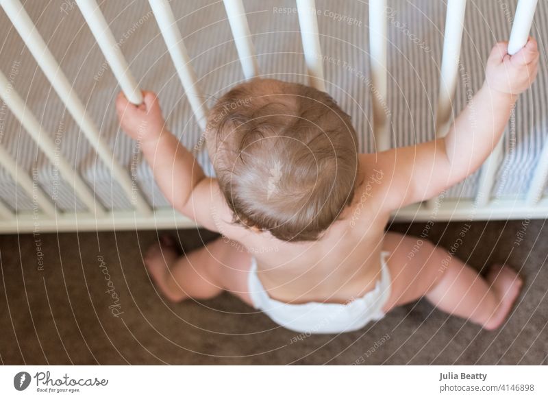 Blick auf ein Kleinkind, das sich zum Sitzen absenkt, während es sich an den Gitterstäben des Kinderbettes festhält; Baby, das lernt, selbstständig zu stehen und zu sitzen