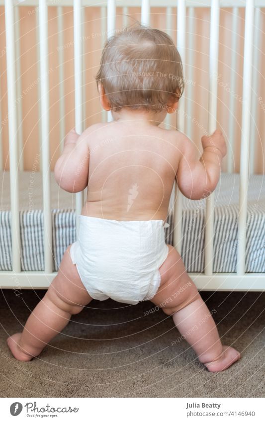 Kleinkind steht mit gespreizten Beinen und hält sich an den Krippenstäben fest: Kinderzimmer mit pfirsichfarbenem, grauem und weißem Dekor Baby Säugling