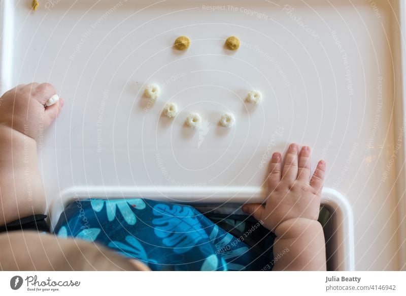 Baby-geführte Entwöhnung macht Spaß: Smiley-Gesicht aus Getreidebällchen auf einem Hochstuhltablett mit der Hand des Babys in der Nähe Säugling Kind