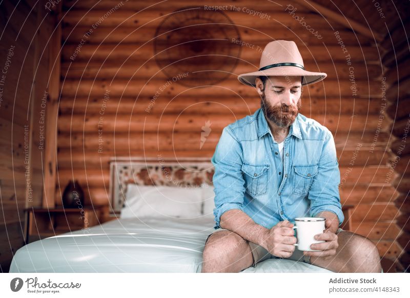 Bärtiger Mann auf Bett in Holzhaus frisch Morgen Kaffee hölzern Haus Reisender rustikal Komfort männlich ruhen sitzen gemütlich heimwärts genießen