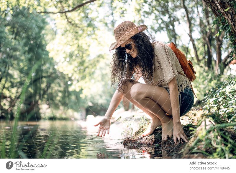 Reisende Frau in der Nähe von Fluss im Wald reisen Tourist Hut Sommer Urlaub Sonnenbrille Wasser Feiertag Gelassenheit Windstille Natur Stein Ufer Wochenende