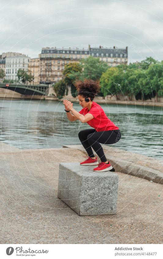 Ethnische Sportlerin hockt beim Training in der Nähe eines Teichs auf einem Stein Kniebeuge Headset Aufwärmen Übung Gleichgewicht Fluss Großstadt