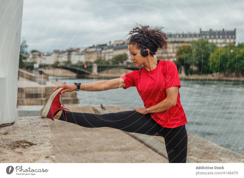 Ethnische Sportlerin mit Headset streckt ihr Bein in der Nähe eines Teiches Athlet Dehnung intelligente Uhr Übung Aufwärmen beweglich Training Fluss benutzend