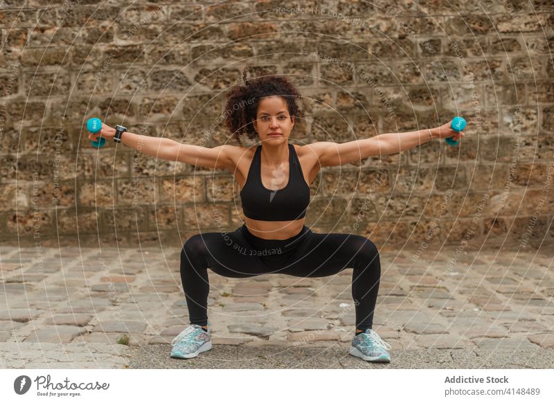 Fitte ethnische Sportlerin in der Hocke mit Hanteln auf einer Böschung während des Workouts Kniebeuge Kurzhantel Training Gleichgewicht erreicht Arme