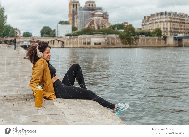 Ethnische Sportlerin mit drahtlosem Headset, die in der Pause über den Fluss nachdenkt nachdenken Drahtlos See zuhören Musik genießen Großstadt Lächeln Wasser
