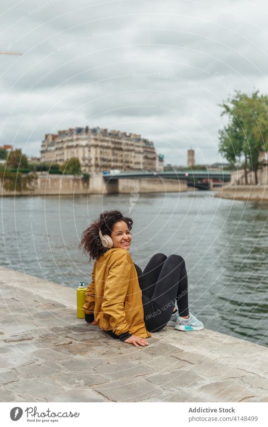 Ethnische Sportlerin mit drahtlosem Headset, die in der Pause über den Fluss nachdenkt nachdenken Drahtlos See zuhören Musik genießen Großstadt Lächeln Wasser